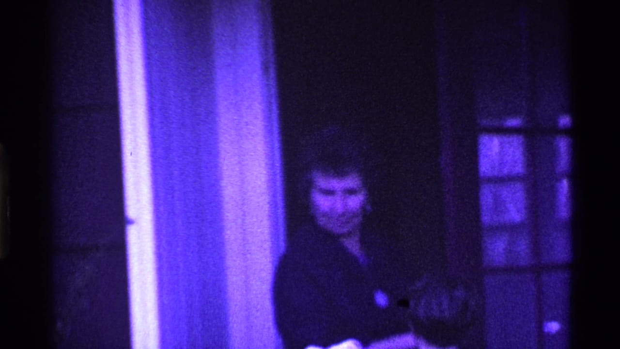 Adult woman stands near a glass-paneled doorway in purple-tinted 8mm footage.