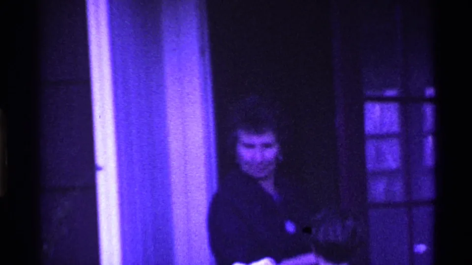 In 1969 Ohio home-movie footage, an adult woman in a dark coat stands near a glass-paneled doorway under strong purple-blue tint.