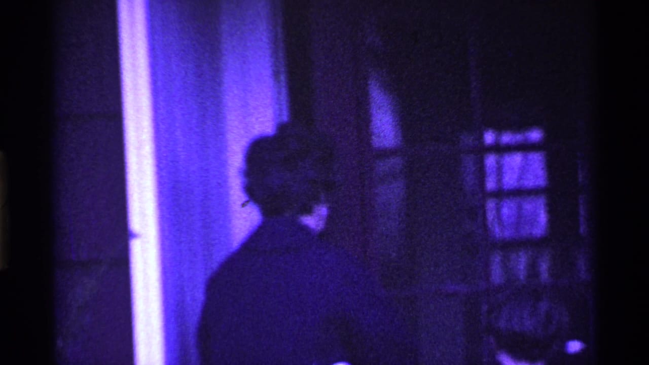 Adult figure turns away near a glass-paneled doorway with a second head low in frame.