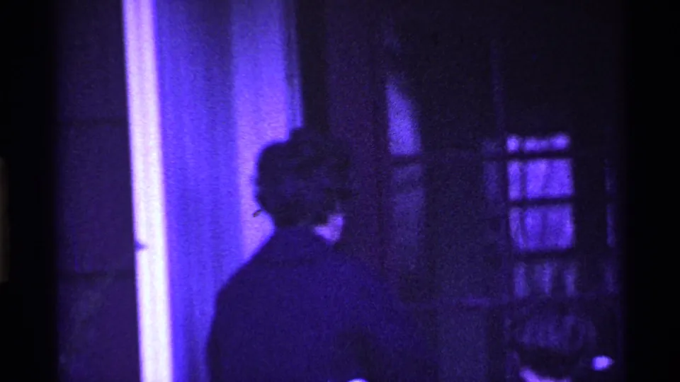 An adult figure turns partly away beside the glass-paneled doorway in purple-tinted 1969 Ohio home-movie footage.