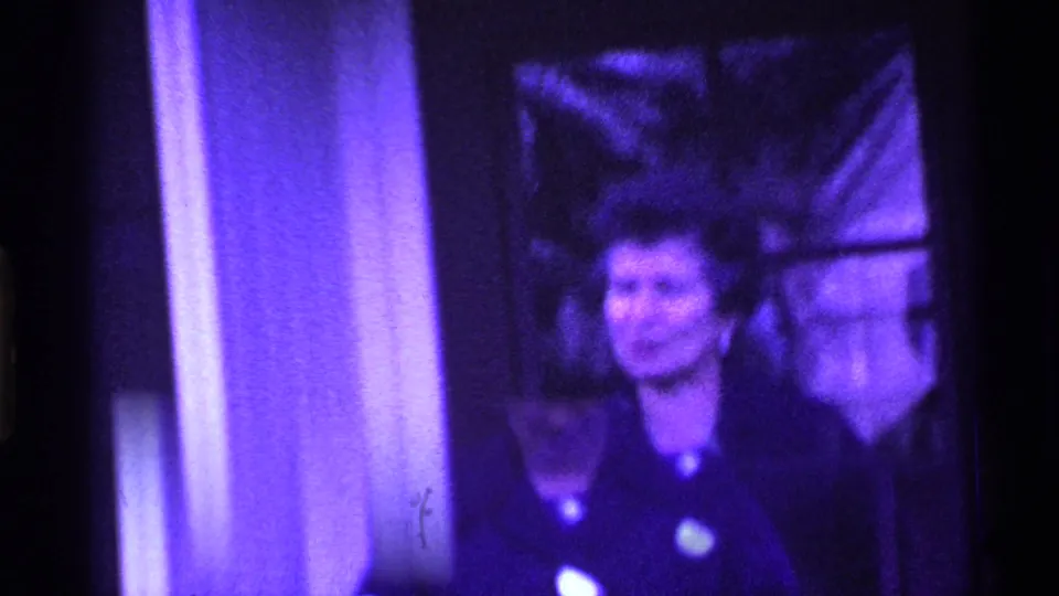 An adult woman in a dark coat faces the camera near the doorway, with another person partly visible behind her in 1969 Ohio 8mm footage.