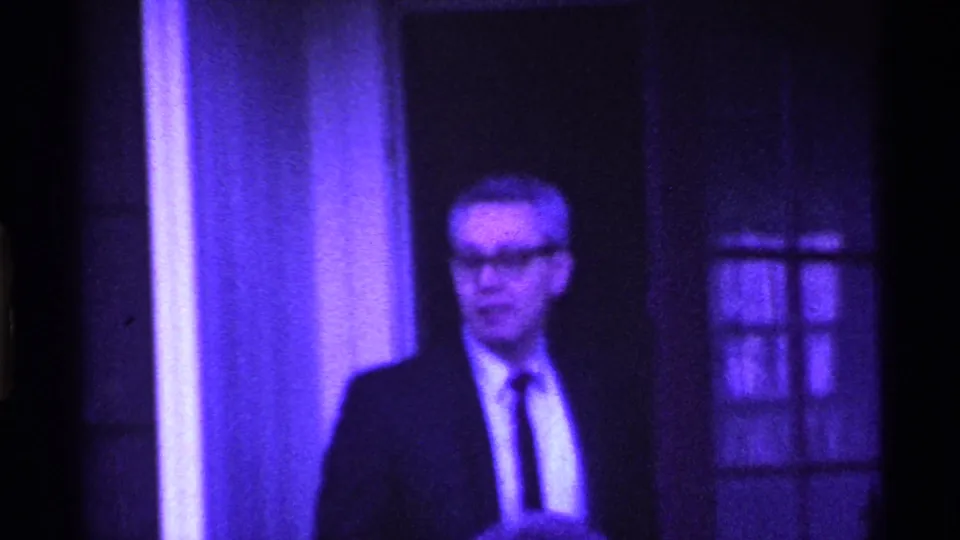 An adult man wearing glasses, a suit, and a tie stands beside the glass-paneled doorway in purple-tinted 1969 Ohio home-movie footage.