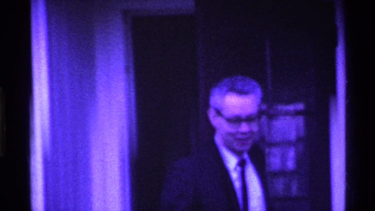 Man in glasses and a suit stands near a doorway under a strong purple color cast.