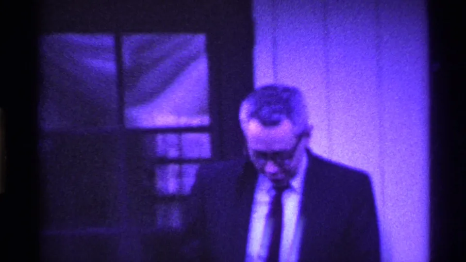 The suited man with glasses looks down beside a dark door and bright wall in the purple-tinted 1969 Ohio 8mm footage.