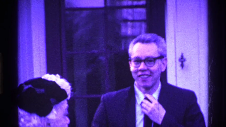 A suited man with glasses smiles and touches his tie, with a partially visible woman at the edge of the 1969 Ohio home-movie frame.