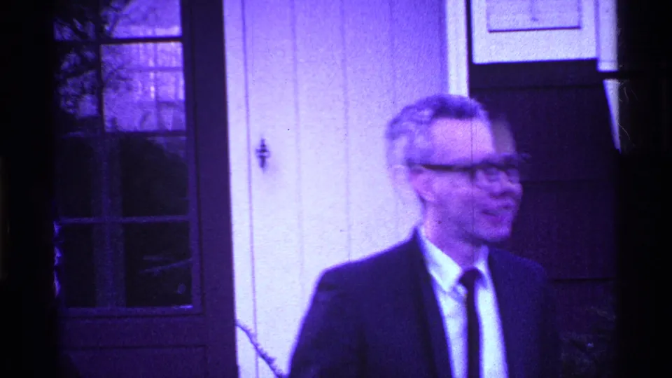 The man in a dark suit and glasses turns sideways outside the doorway, with window panes and a light wall visible in the Ohio footage.