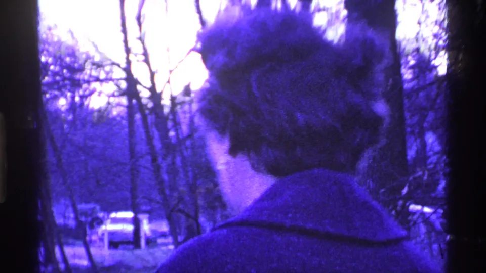 1969 Ohio home-movie frame of a woman in a dark coat walking away through a wooded area, with cars faintly visible beyond the trees.