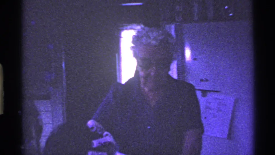 1969 Ohio home-movie frame of an older woman working near cabinets in a private kitchen or dining area.