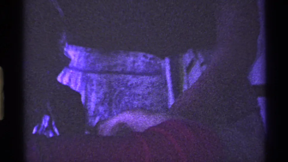 Grainy 8mm frame from the Ohio home movie showing a close, low-light view of clothing and upholstery in a purple-lit room.
