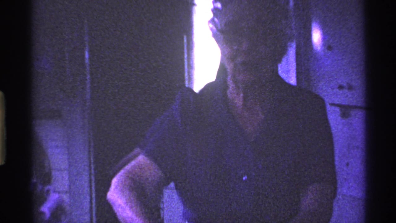 A woman in glasses, dark shirt, and light apron stands in a dim Ohio kitchen in this 1969 home-movie frame.