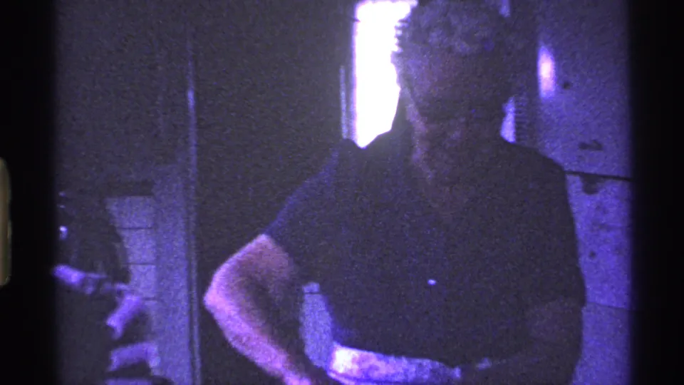 The woman, wearing glasses and a light apron over a dark shirt, stands in a private Ohio kitchen during the 1969 home-movie scene.