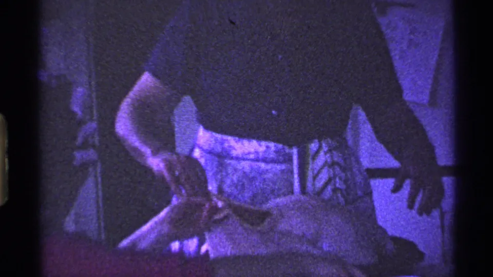 A woman in a light apron reaches toward a turkey on the counter in a dim 1969 Ohio kitchen home-movie frame.
