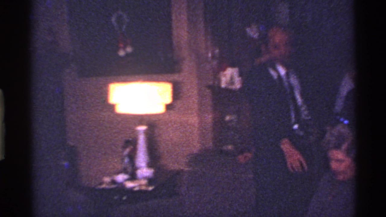 Dim living room with table lamp, seated adults, and dark formal clothing.