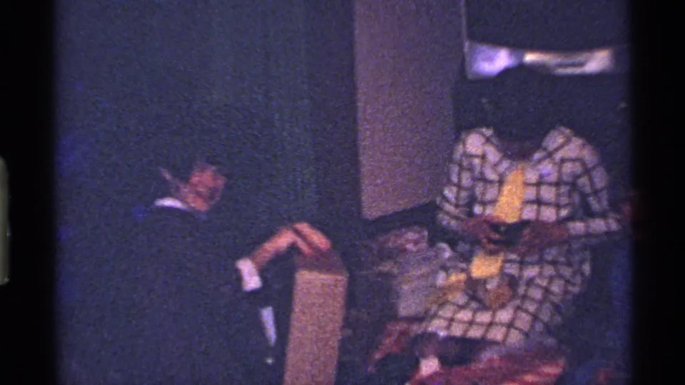 1969 Ohio home-movie frame with two adults seated indoors, including one person in a bold patterned jacket.