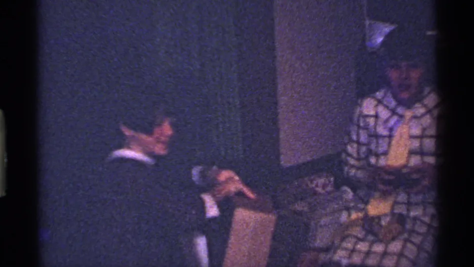 In a dim Ohio home interior from the 1969 8mm clip, one child handles a gift box as another child in plaid looks on.
