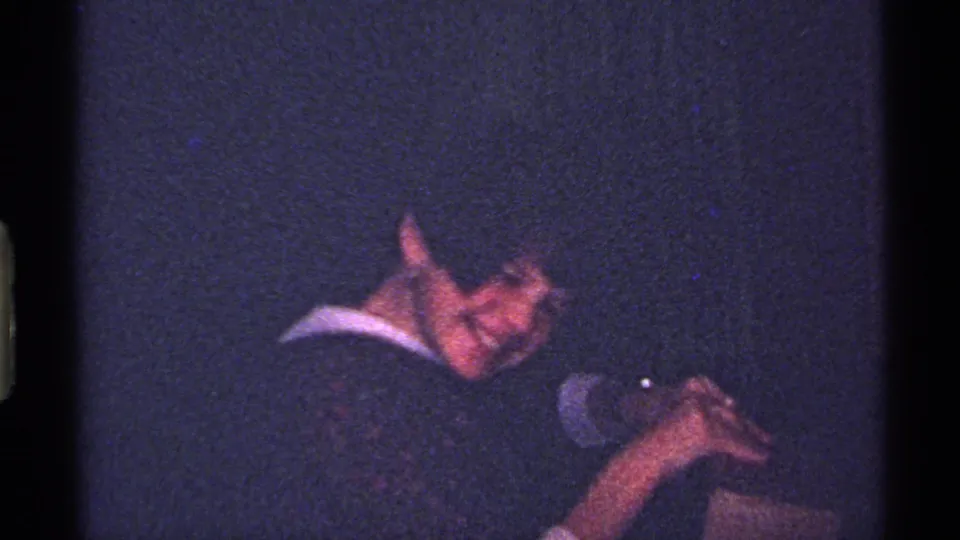 A child in a dark suit and white collar leans back with a small present in a dim 1969 Ohio home-movie frame.