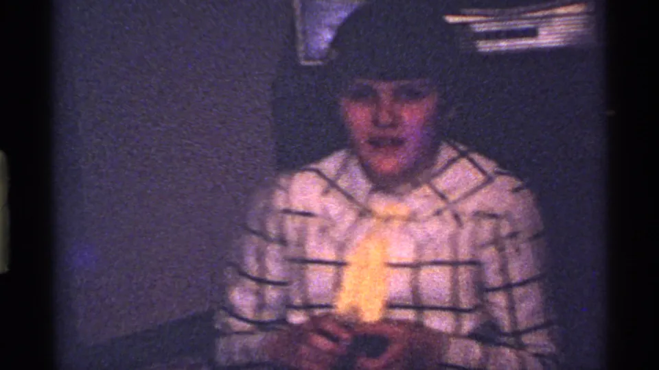 A child in a plaid shirt and yellow tie faces the camera in a low-light Ohio home interior from the 1969 8mm footage.