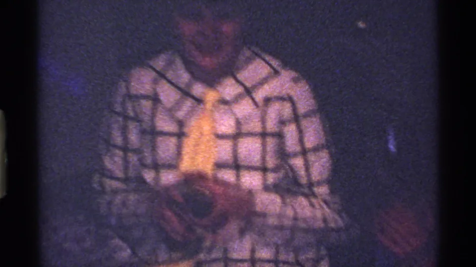 The child in plaid and a yellow tie looks down at an object in the dim private interior of the 1969 Ohio home movie.