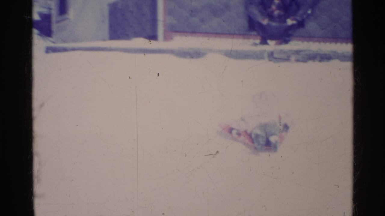 Bundled person lying on a sled on a snowy slope, with faint residential background at the top of frame.