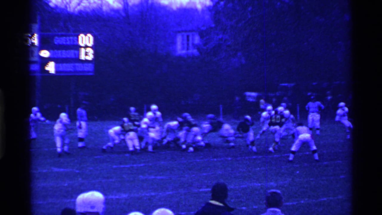 Football players line up on a field with a scoreboard and spectators visible.