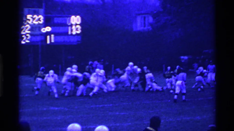 A tackle develops during the Ohio high school football game, with helmeted players clustered under the blue cast of 8mm night footage.