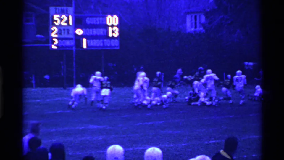 Players regroup after a 1969 Ohio high school football play, with a scoreboard, sideline spectators, and school grounds visible in the frame.