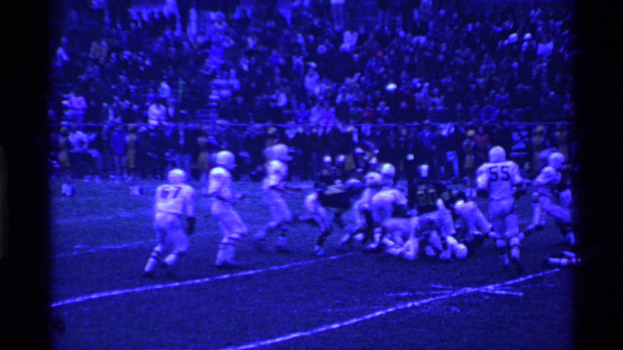 Football play on a grass field with white-uniformed players, a referee, and crowded bleachers behind them.