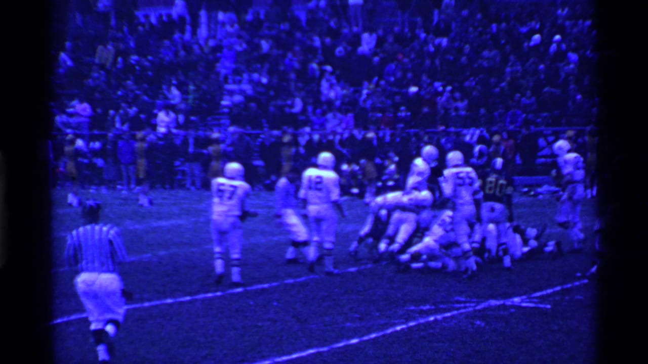 Blue-tinted 8mm frame of a football game in Ohio, with a referee moving toward a pile of players and a stadium crowd in the background.