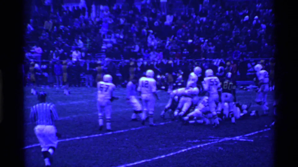 Blue-tinted 8mm frame of a football game in Ohio, with a referee moving toward a pile of players and a stadium crowd in the background.