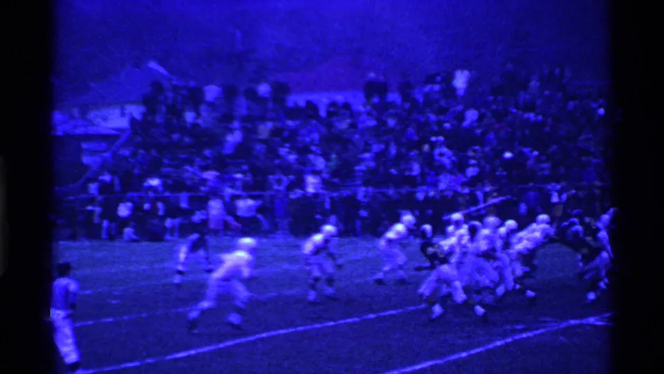 Wide 1969 Ohio football frame from a home movie, showing players in motion on a grass field below packed small-stadium bleachers.