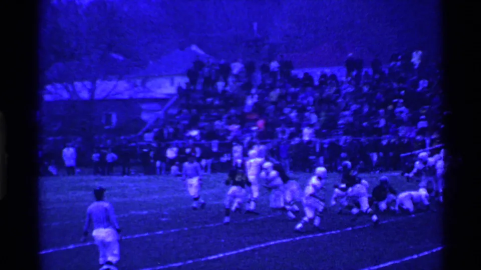 Home-movie frame of a 1969 Ohio football game, with a striped referee in front of players and a crowd watching from the bleachers.