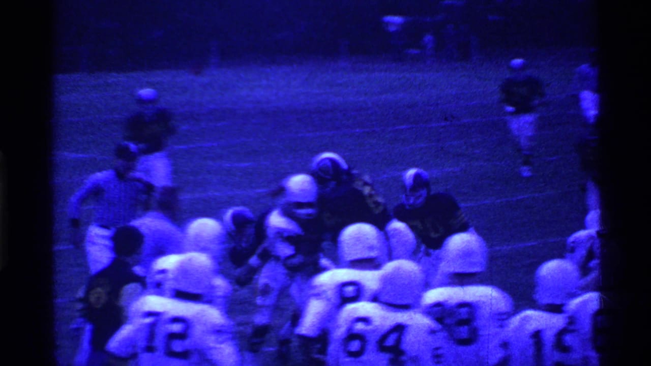 Football players in white and dark uniforms converge near the sideline on a blue-tinted field.