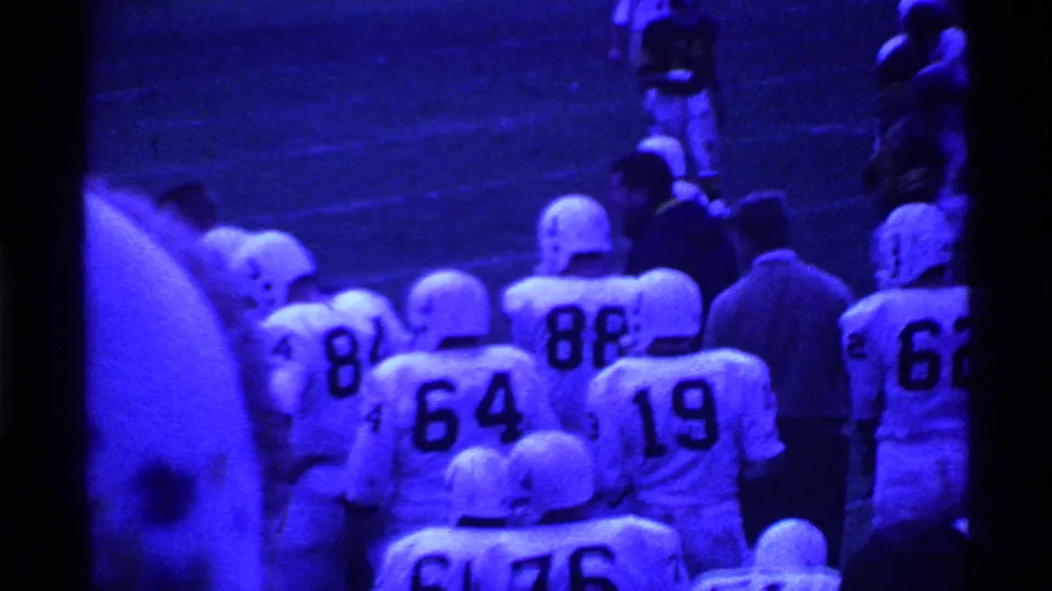 Blue-shifted 1969 Ohio 8mm footage shows helmeted players in numbered white jerseys waiting along the football sideline.