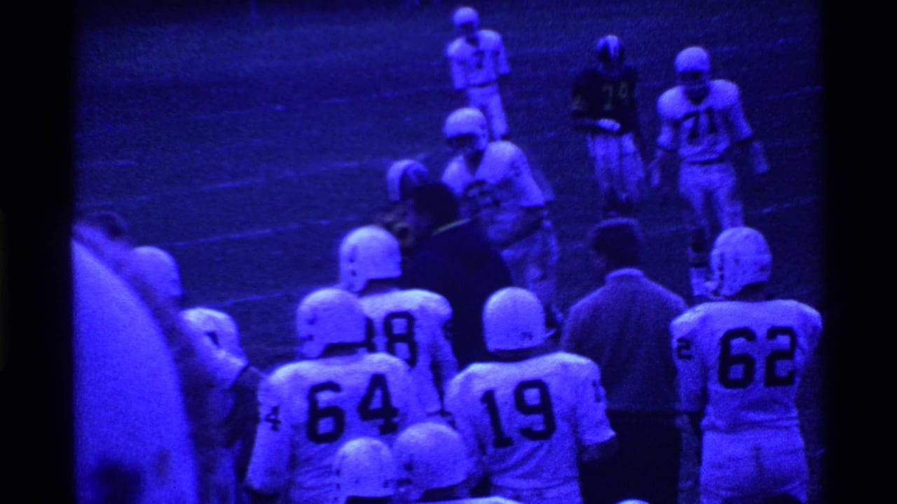Helmeted football players gather near the sideline on a blue-tinted field.
