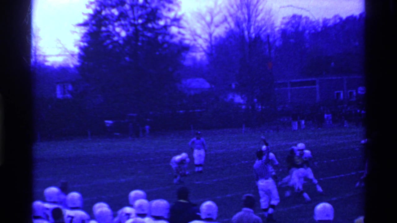 Football players line up on a field with trees and houses in the background.