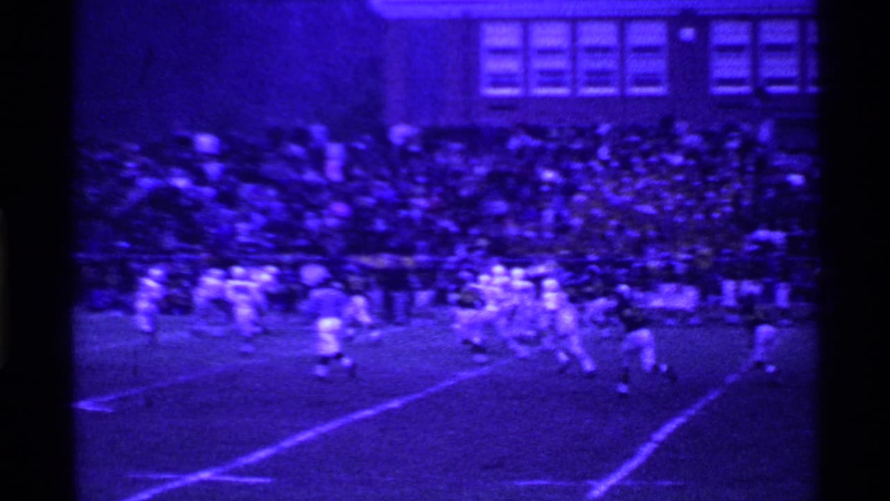 Football players line up on a field with crowded bleachers behind them.