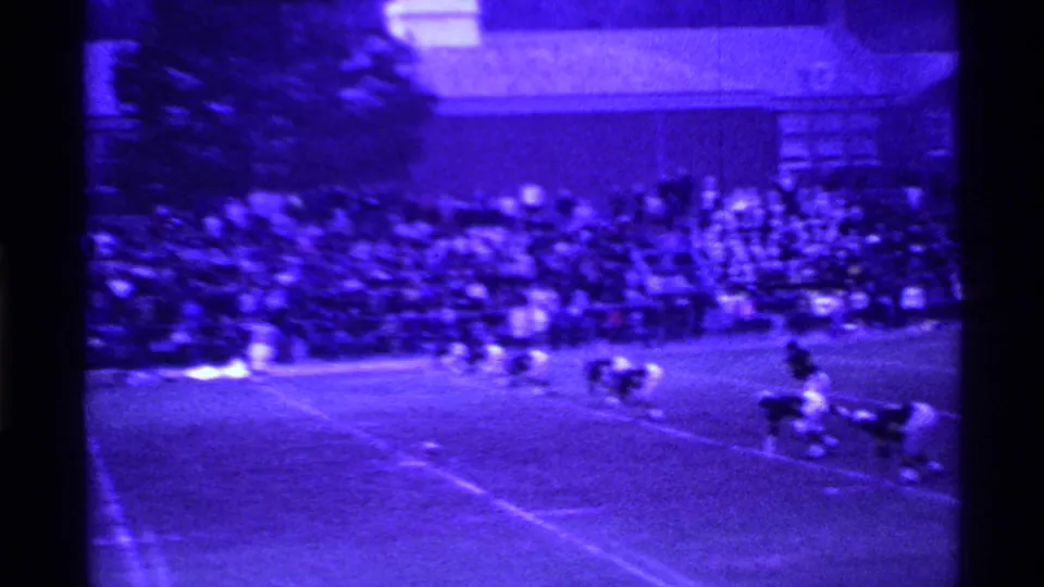 Wide purple-tinted 8mm frame of a 1969 Ohio high school football field with players and spectators visible.