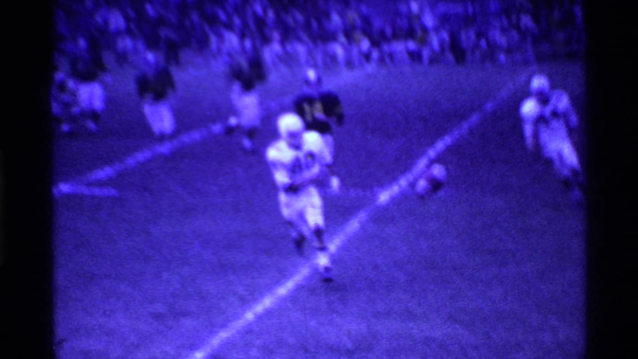 Football players on a field during a night game, with bleachers visible behind them.