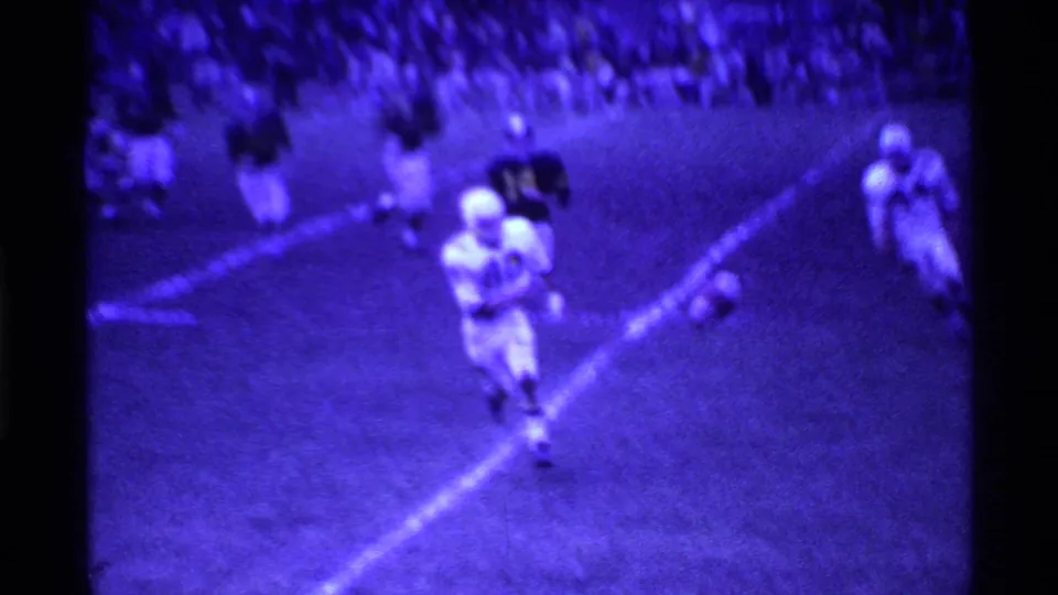 1969 Ohio home-movie frame showing football players in white uniforms on a grass field under purple-tinted night lighting.