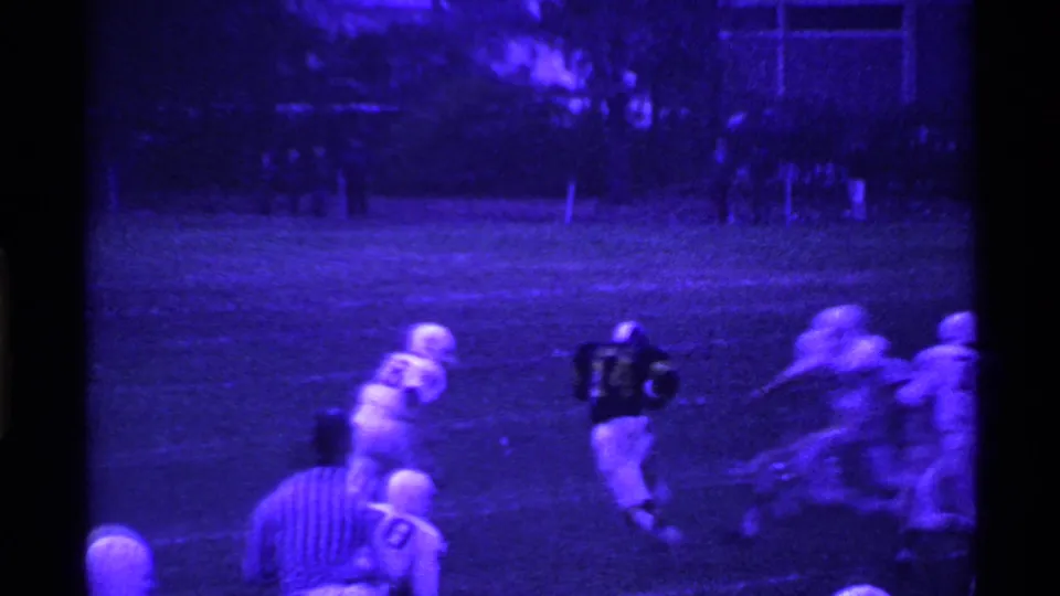 Wide purple-tinted 8mm frame of a 1969 Ohio night football play with players in white and dark uniforms.