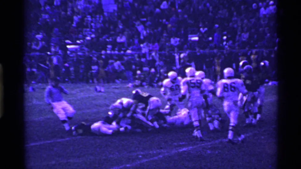 1969 Ohio 8mm frame showing helmeted football players around a tackle on a grass field, with crowded stands in the background.