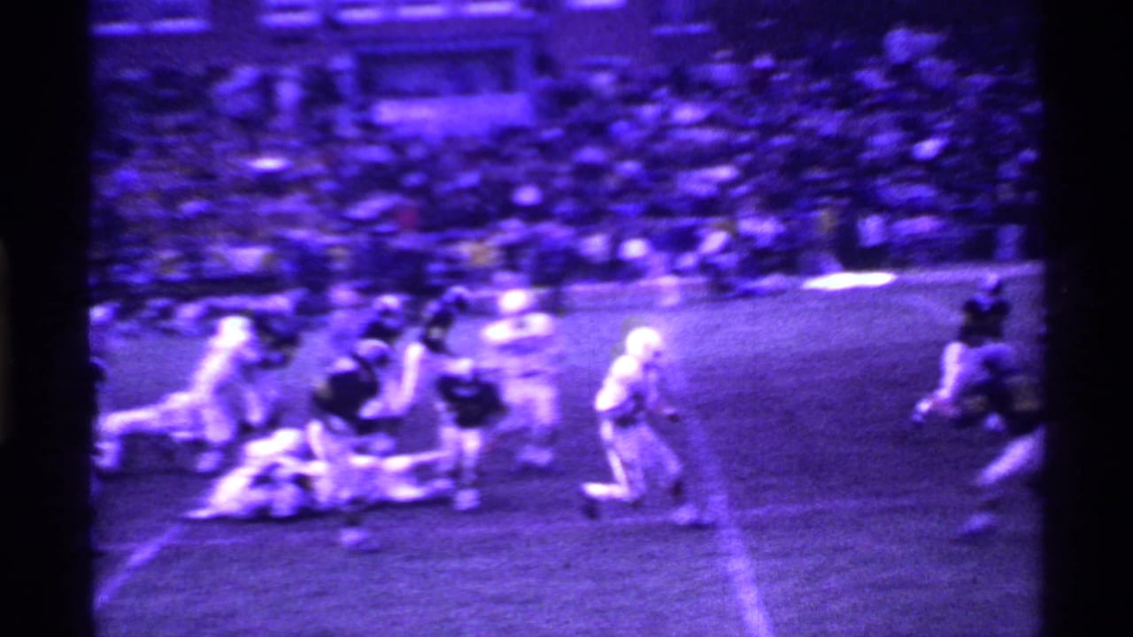 Football play continues near a yard line, with players falling and a crowded stand behind the field.