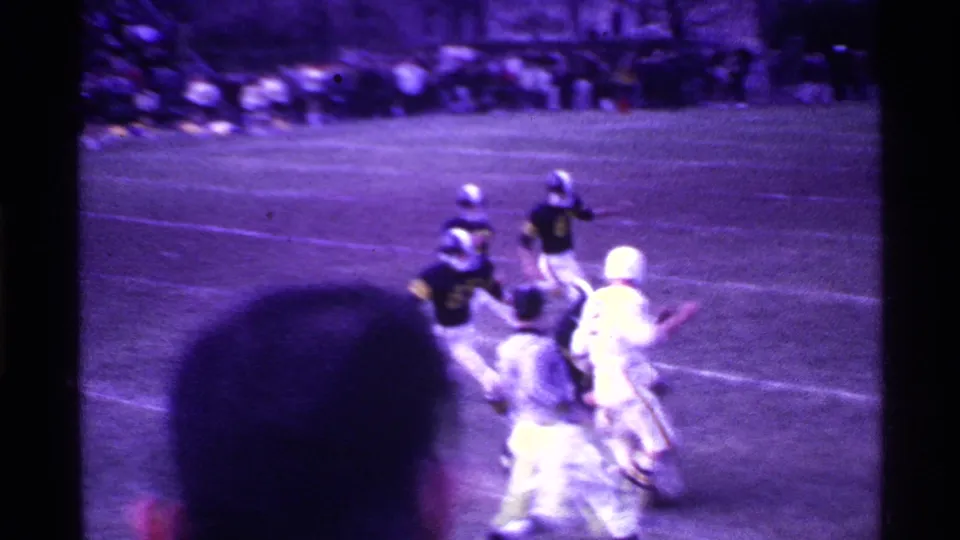 1969 Ohio home-movie frame of a football play moving across the field, with players in dark and white uniforms and a foreground spectator head.