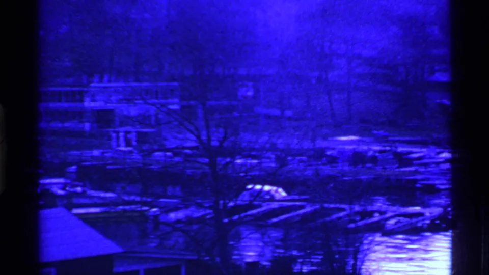 Ohio 1969 home-movie frame of a blue waterfront settlement with moored boats, docks, bare trees, and low houses across reflective water.