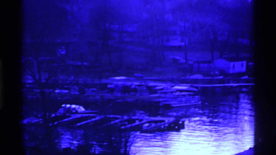 Ohio 1969 8mm frame with a softer blue view of lakeside docks, small boats, bare trees, shoreline buildings, and reflected light on water.