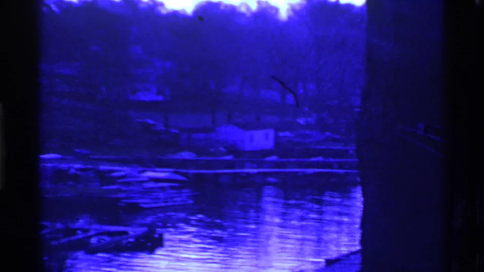 Ohio 1969 home-movie frame showing reflective water, docks, small boats, bare trees, and a small shoreline structure in deep blue monochrome.