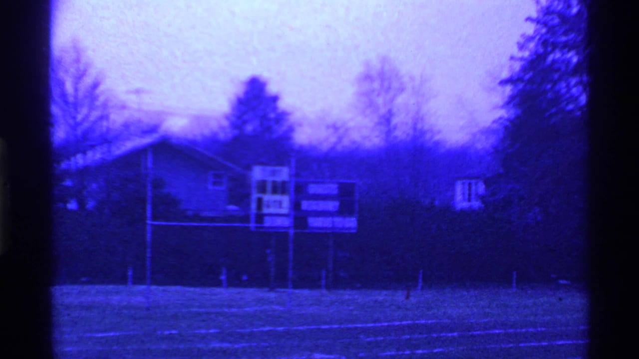 Blue-tinted view of an empty football field with a scoreboard, trees and buildings beyond.