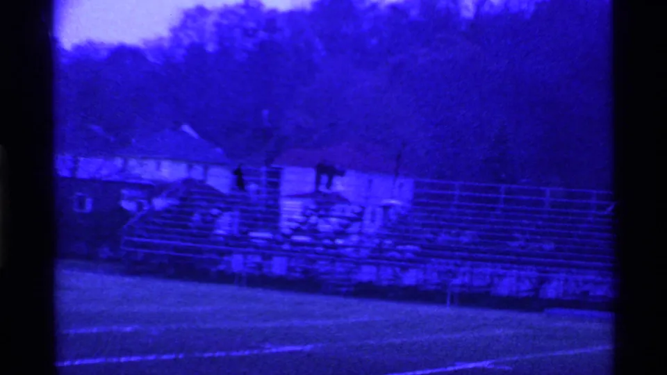 Soft 1969 Ohio 8mm view of empty stadium bleachers, field markings, rooftops and trees under a strong blue cast.