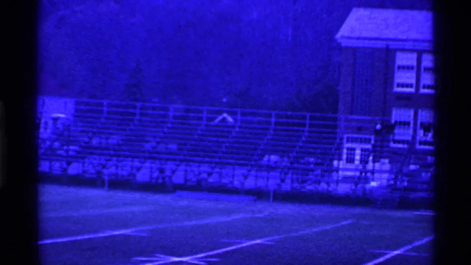 Blue-shifted 8mm frame showing empty bleachers, field markings, trees and a brick school-like building in Ohio.