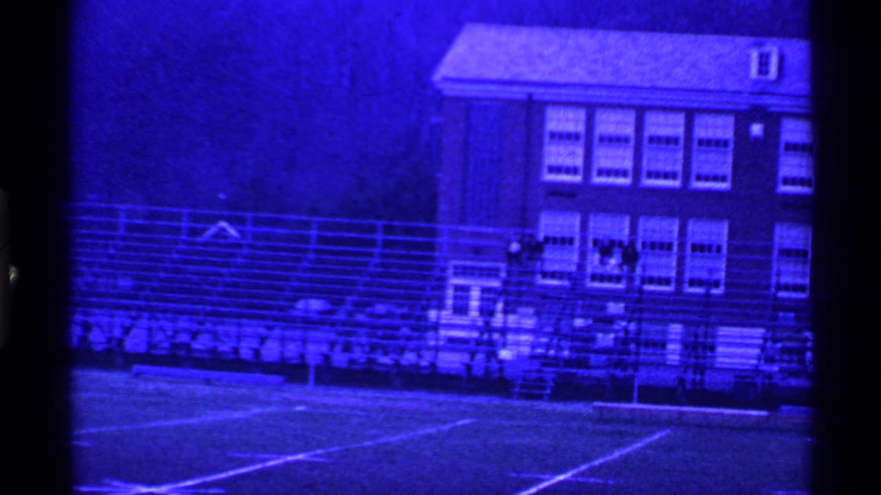 1969 Ohio 8mm frame showing a blue-tinted school sports field with bleachers and a brick building behind it.